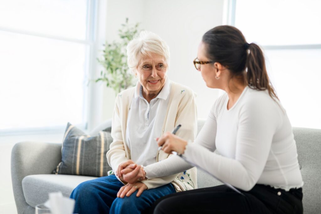 Eine Pflegeberaterin führt ein Gespräch mit einer Seniorin. Beide sitzen auf dem Sofa.